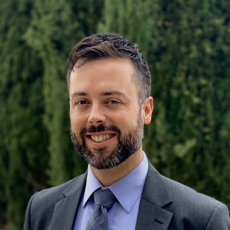 A headshot of Adam, smiling into the camera with short brown hair and a beard, wearing a grey and blue suit.