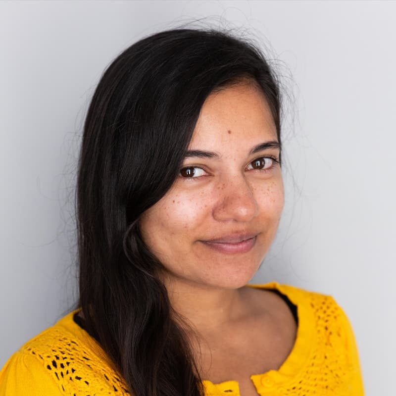 A headshot of Kamla, smiling at the camera with long black hair and a yellow sweater.