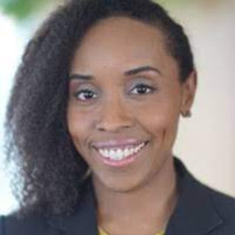 A headshot of Nakia smiling at the camera with black curly hair and black suit