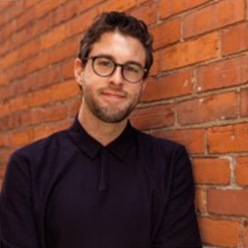 A headshot of Tyler, with a beard and short brown hair and dark-rimmed glasses smiling at the camera