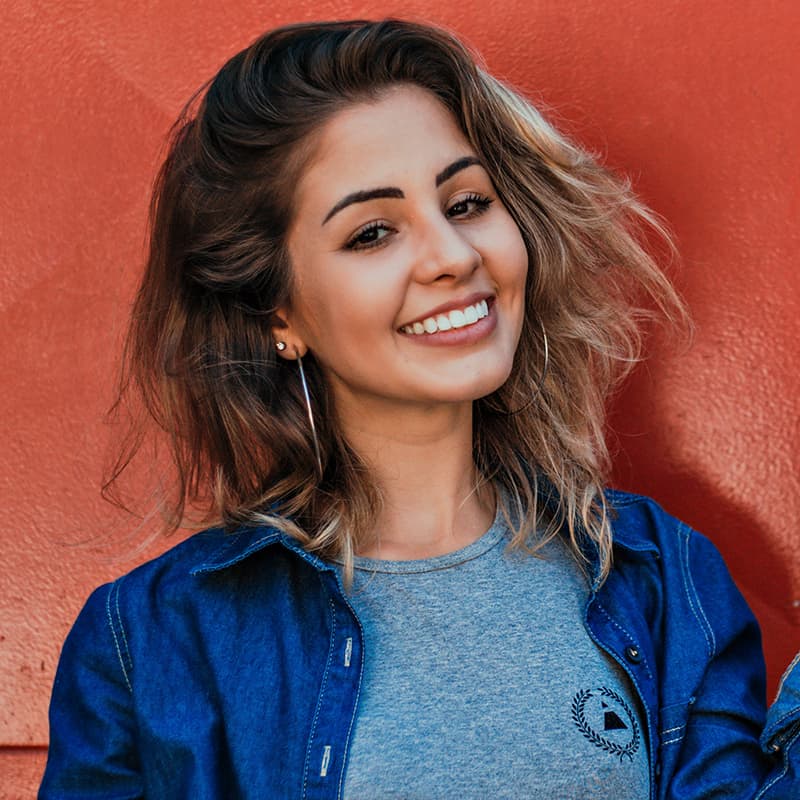 A young person with long brown hair, hoop earrings, and a jean jacket, smiling at the camera in front of a rusty coloured wall.