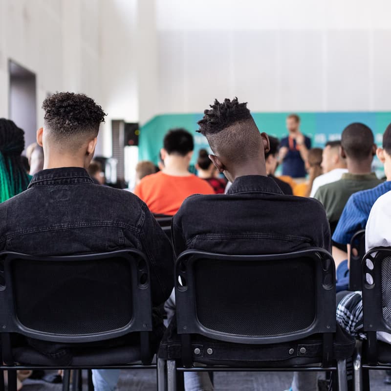 Students of various genders and ethnicities sit in a large room, attending a presentation. The viewpoint is from the back of the room.