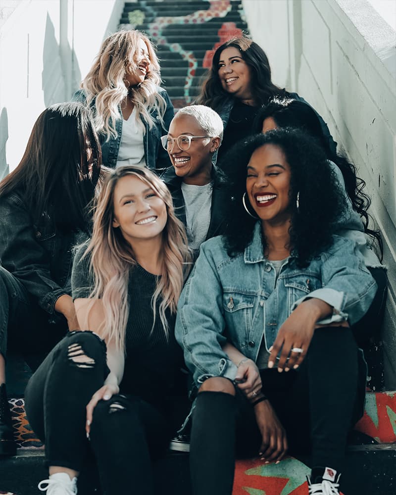 A diverse group of seven young folks, early twenties, sitting on a stairway and smiling and laughing.