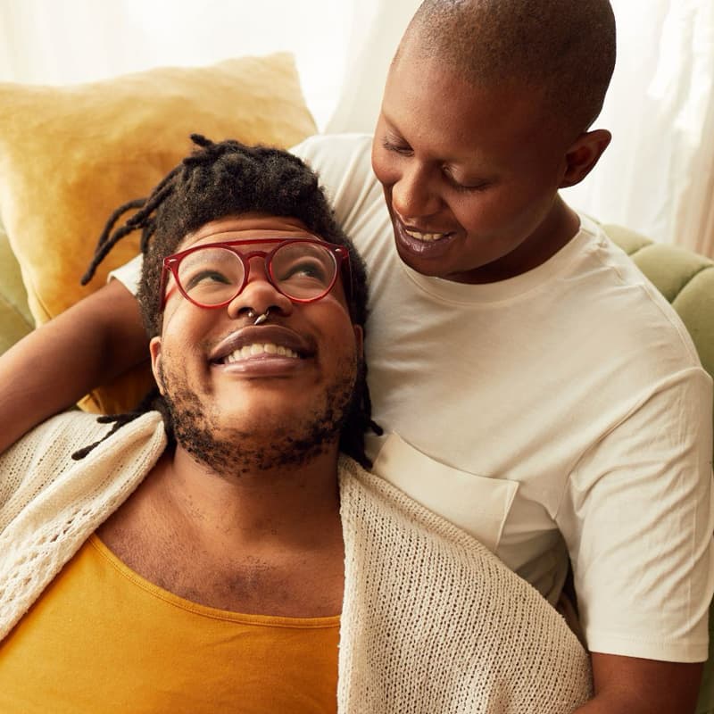 Two young Black folks cuddling on a green couch and smiling at one another.