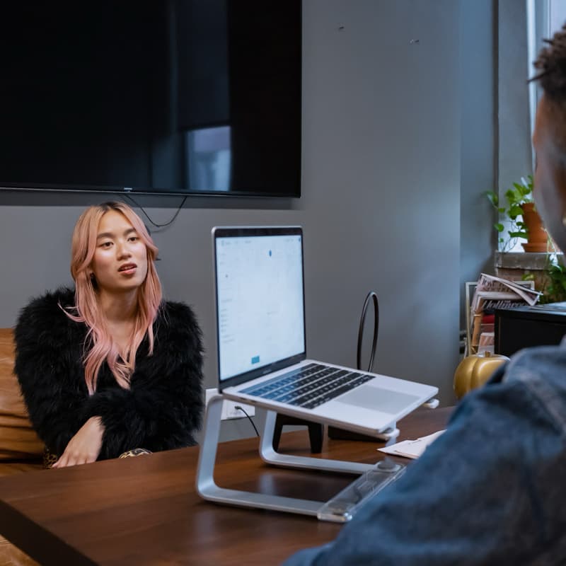 Two young people sit in an office. A transfeminine person with pink hair faces the camera while sitting on a leather couch. A person with short black hair has their back to the camera and sits in front of a laptop. They are wearing a jean jacket and earrings.