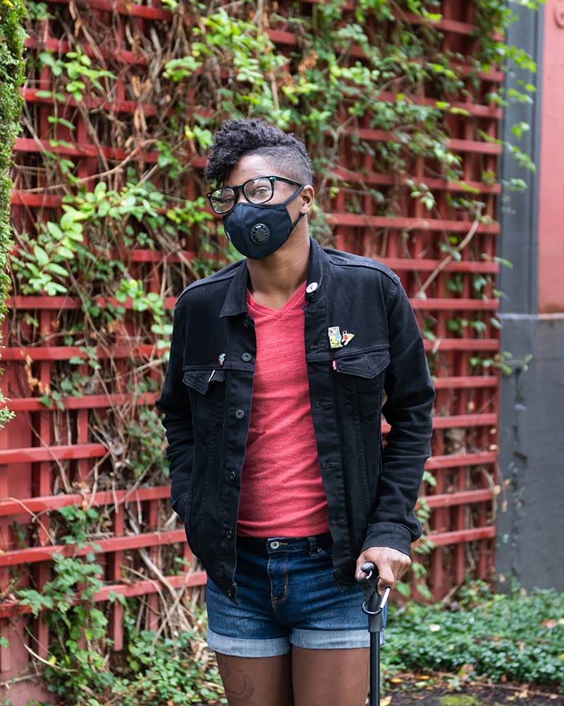 A Black person wearing a face mask, a red t-shirt, and holding a cane, standing outside of a red trellised wall.